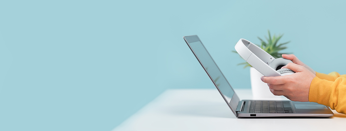 Side view of slim laptop and hands with wireless headphones on grey desk. Blue background.