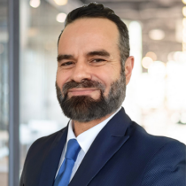 Man with dark hair and beard with a white shirt, blue tie and navy blazer.